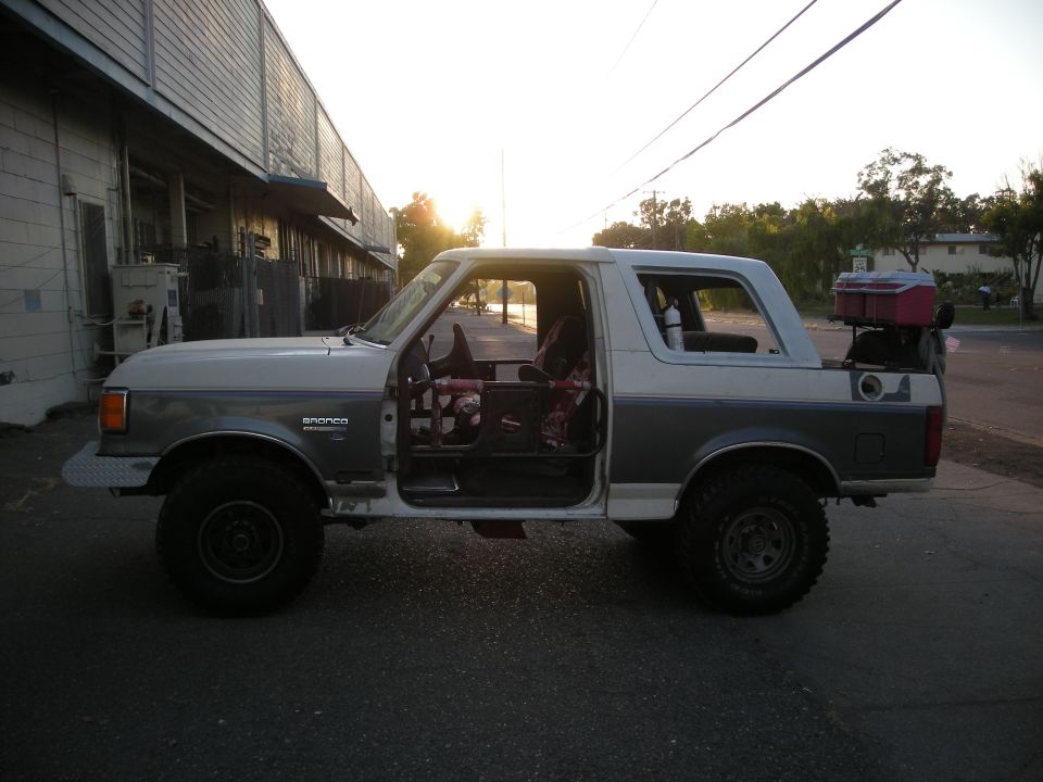 Side view of a 1991 Ford Bronco with doors removed, parked outside a building at sunset