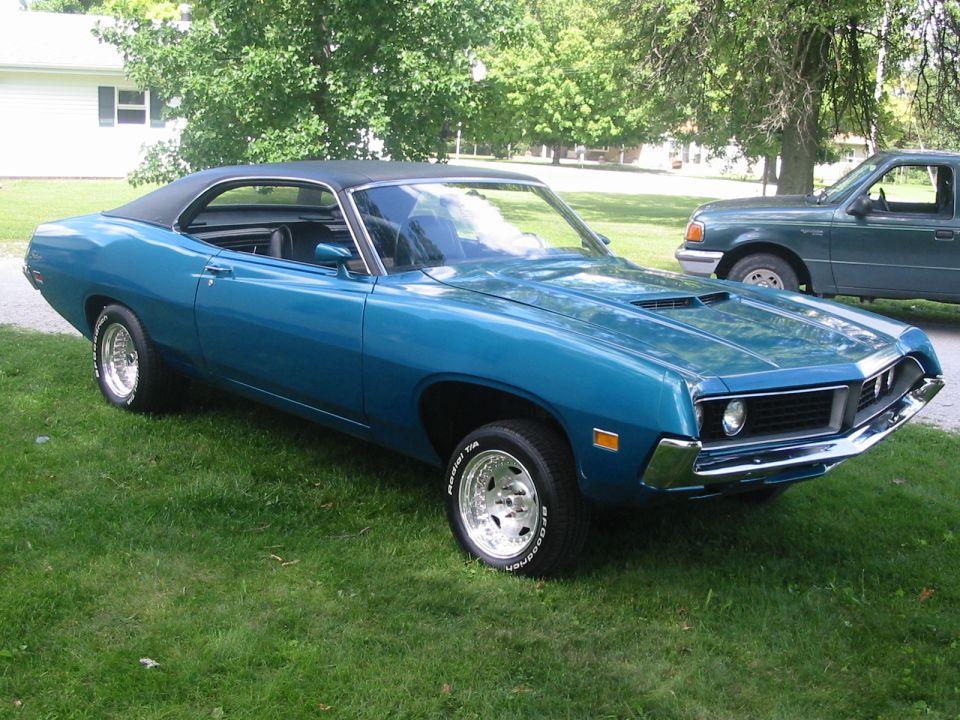 Blue 1970 Ford Fairlane coupe with black vinyl roof parked on grass, green pickup truck behind it