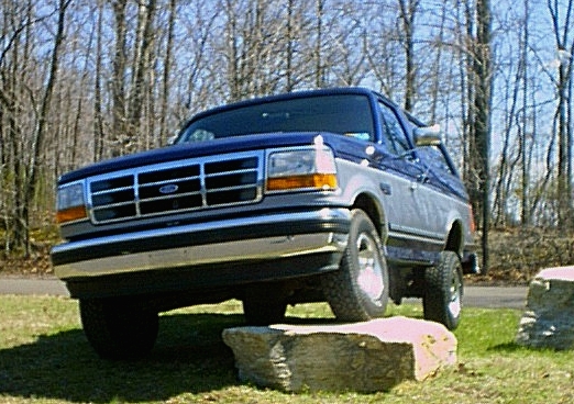 Dark blue Ford Bronco SUV seen from a low front angle on grass with rocks in foreground and bare trees behind.