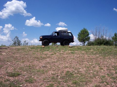 1996 Ford Bronco - Driver-side profile of a lifted dark blue full-size Ford Bronco parked on a rocky, sparsely grassed ridge under blue sky with puffy clouds.…