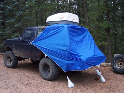 1996 Ford Bronco - Muddy dark blue lifted Ford Bronco at a pine-forest campsite with a large blue tarp rigged off the rear as an awning, weighted at the corne…