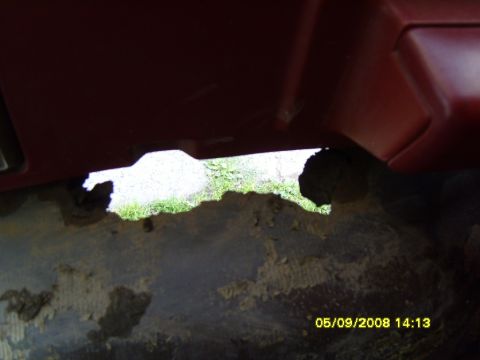 1989 Ford Bronco - Close-up of a dark red body panel on a Ford Bronco showing severe rust perforation, with a large jagged hole revealing grass and pavement b…