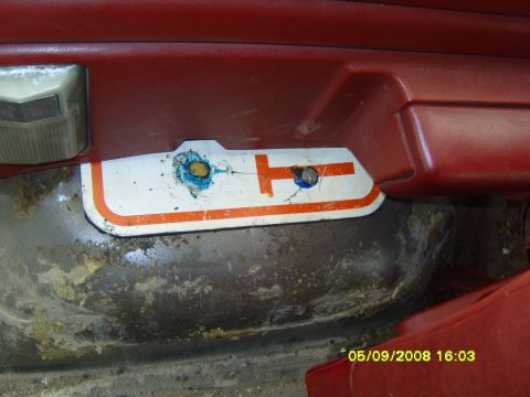 1989 Ford Bronco - Interior detail of a red door panel with the carpet and lower trim removed, exposing bare, surface-rusted sheet metal at the door sill. A w…