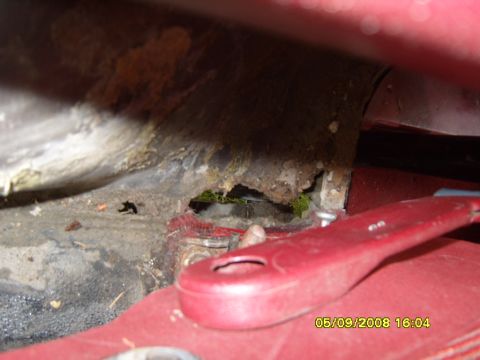 1989 Ford Bronco - Tight close-up of rust-through in a red truck's inner fender/cowl area, with a jagged rot hole opening onto grass below and heavy surface c…