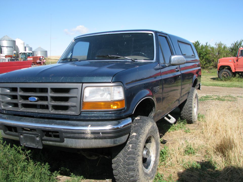 Dark blue Ford Bronco with camper shell parked in a grassy field, farm silos in background.