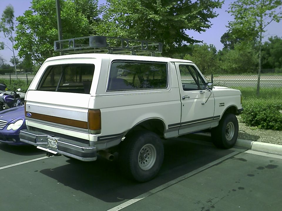 White 1990 Ford Bronco with roof rack, parked next to a blue Mercedes-Benz sedan.