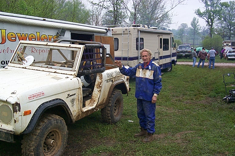 Man in blue jumpsuit next to white modified Ford Bronco with roll cage in a muddy field with RVs.
