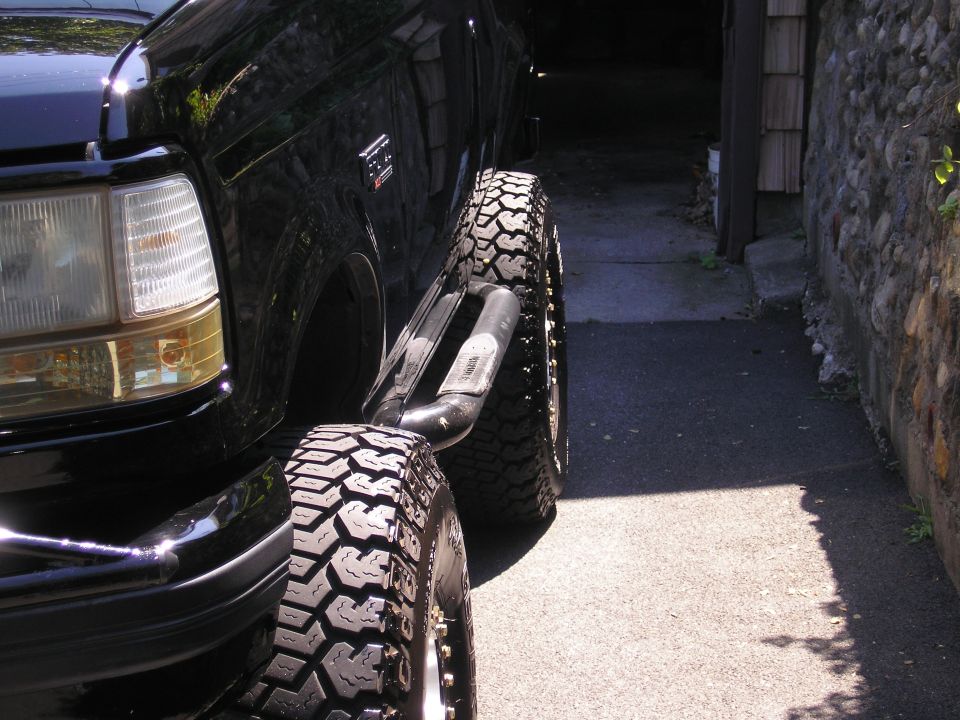 Black Ford Bronco front corner with off-road tire and side step, parked on asphalt next to stone wall.