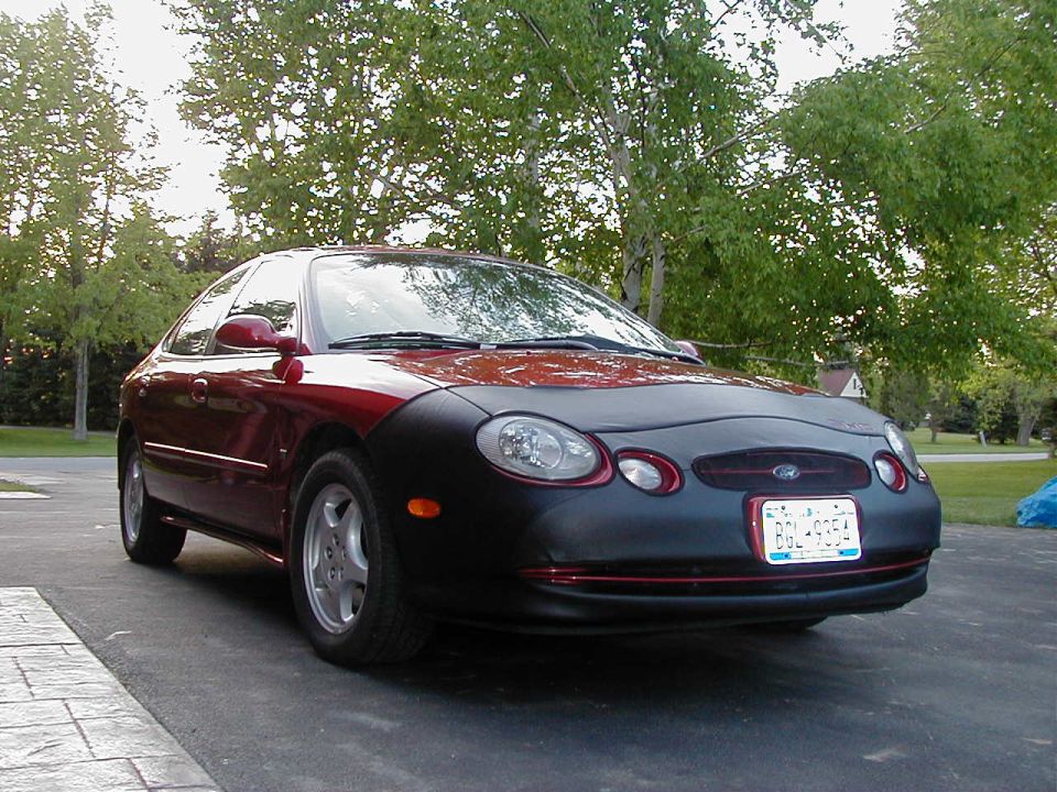 Dark red Ford Taurus sedan with black front-end bra, 3/4 front view on driveway with trees behind