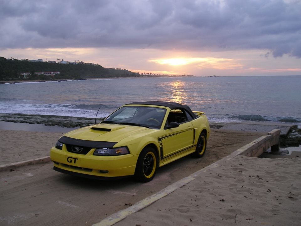 Yellow Ford Mustang convertible parked on a beach at sunset.