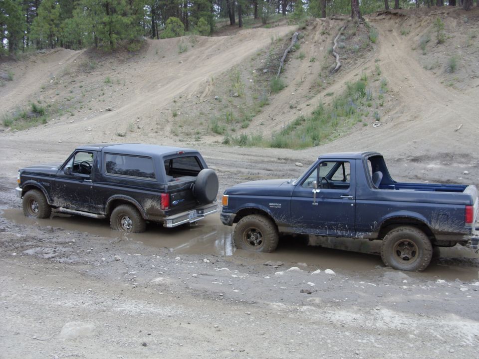 Two Ford Broncos, one black SUV and one blue pickup, stuck in mud on a dirt track.