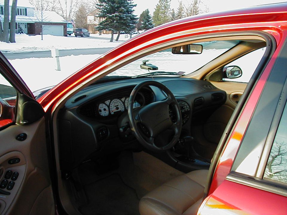 Interior of red sedan through open driver's door, tan leather seats, white gauges, snowy neighborhood outside