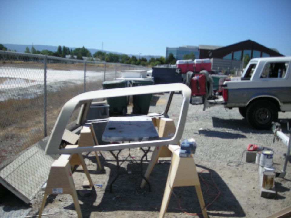 White and gray Ford Bronco parked outdoors, with a white vehicle frame on sawhorses in the foreground.