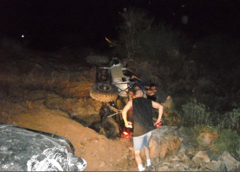 1986 Toyota Hilux - An overturned silver off-road vehicle lies on its side at night, with several people gathered nearby.