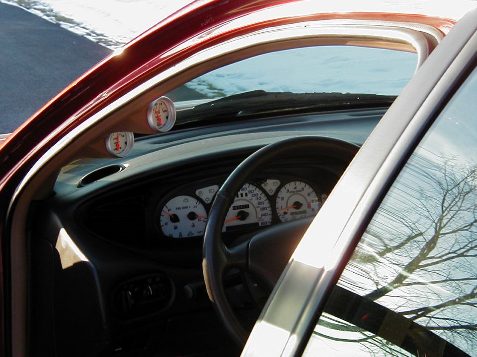 Car interior with two aftermarket gauges mounted on the A-pillar and a dashboard with white-faced gauges. The steering wheel is visible