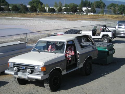 1991 Ford Bronco - A two-tone Ford Bronco with its doors off sits parked next to a dumpster and a white Jeep.