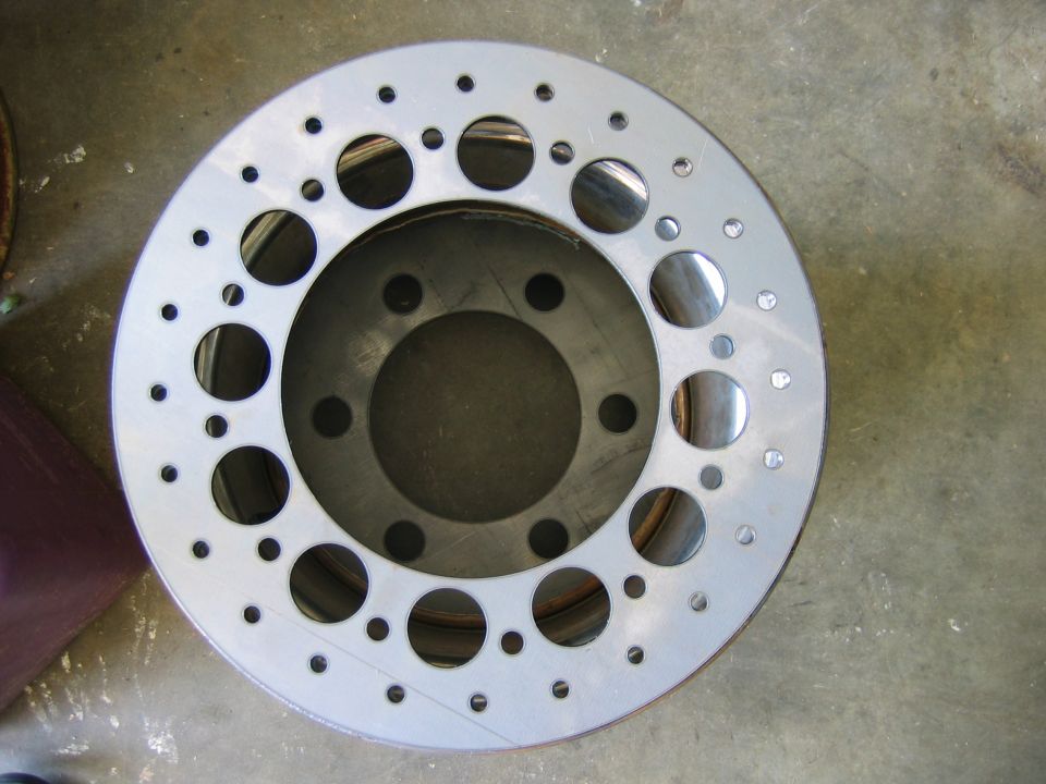 Gray drilled brake rotor with multiple holes of varying sizes, resting on a concrete floor