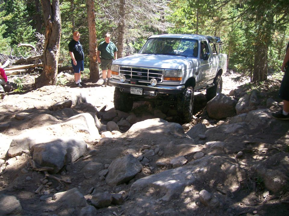 White Ford Bronco driving on a rocky trail in a forest with two men watching.
