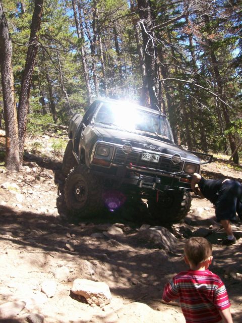 1978 Ford Bronco - A dark-colored, lifted Dodge pickup truck is navigating a rocky, wooded trail. A person is leaning against the front of the truck, and a yo…