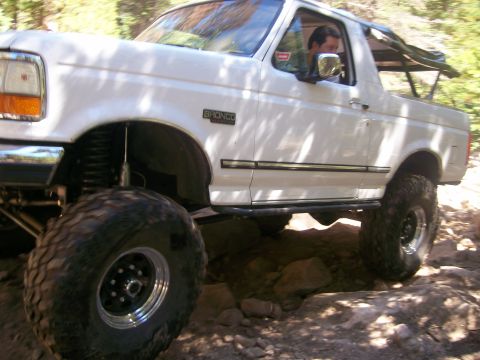 1978 Ford Bronco - The front driver's side of a lifted white Ford Bronco is shown navigating a rocky, off-road trail. The vehicle features large, aggressive t…