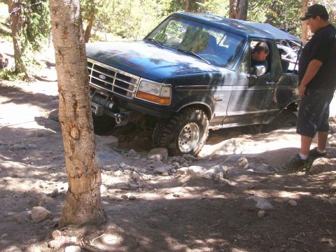 1978 Ford Bronco - A dark blue Ford Bronco is stuck on a rocky trail, with its front tire high on a rock. A man stands next to the vehicle, observing.