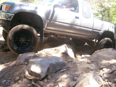 1978 Ford Bronco - A silver pickup truck is navigating a rocky, off-road trail. The front tire is angled up on a large rock, showcasing the vehicle's off-road…