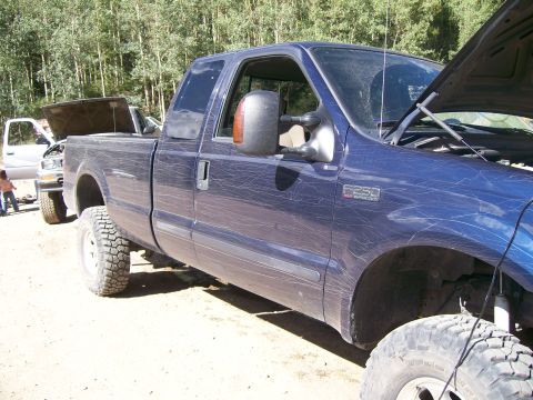 1978 Ford Bronco - A blue Ford F-250 Super Duty pickup truck is parked outdoors on a dirt road, with its hood open. Another white truck with its hood open is…