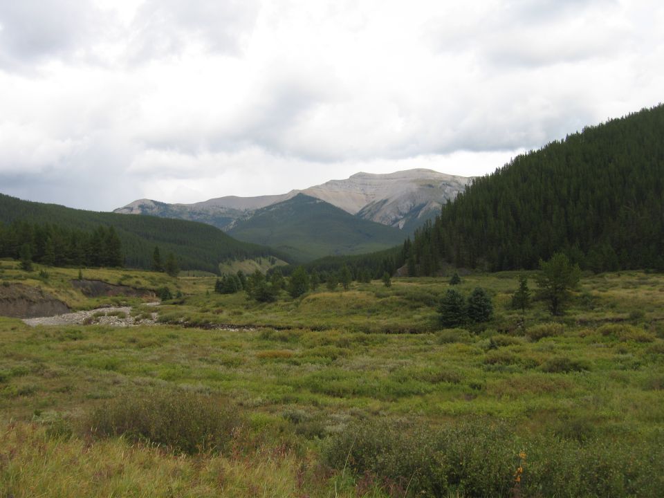 Valley with green hills, mountains, and a cloudy sky.