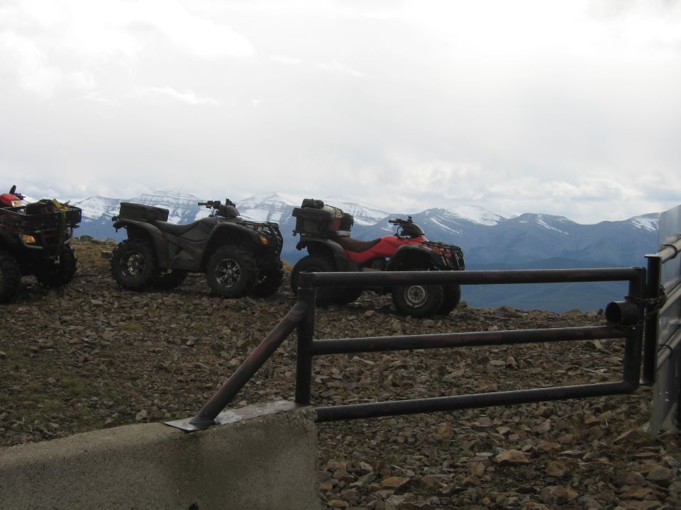 Three ATVs parked on a rocky mountain overlook with snow-capped mountains in the background.