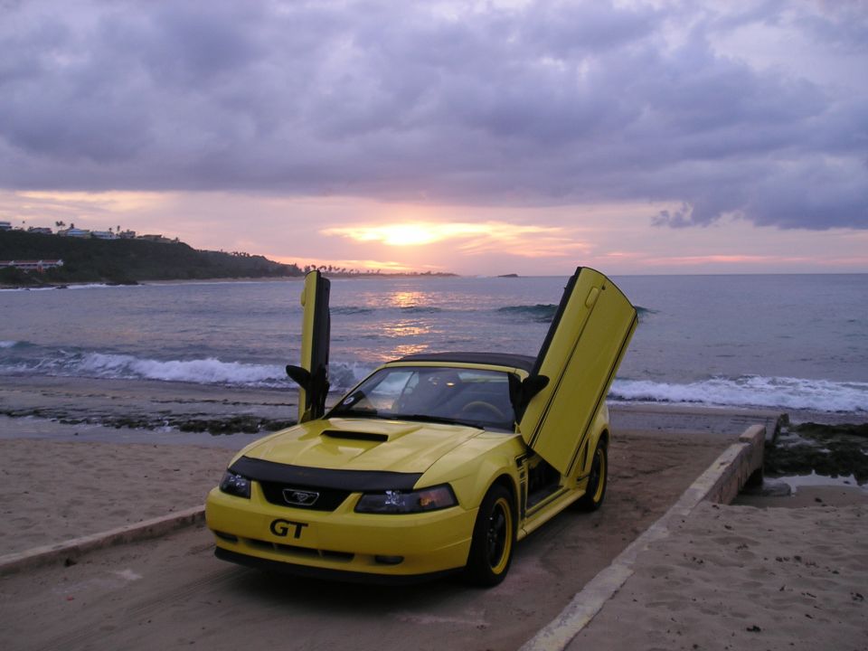 Yellow Ford Mustang convertible with lambo doors open on a beach at sunset.
