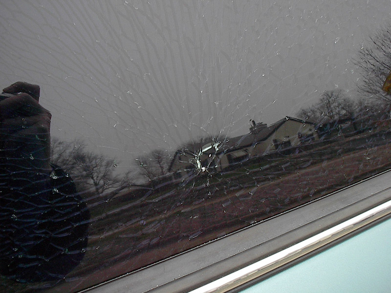 Shattered car window with spiderweb cracks from an impact point, reflecting trees and a house