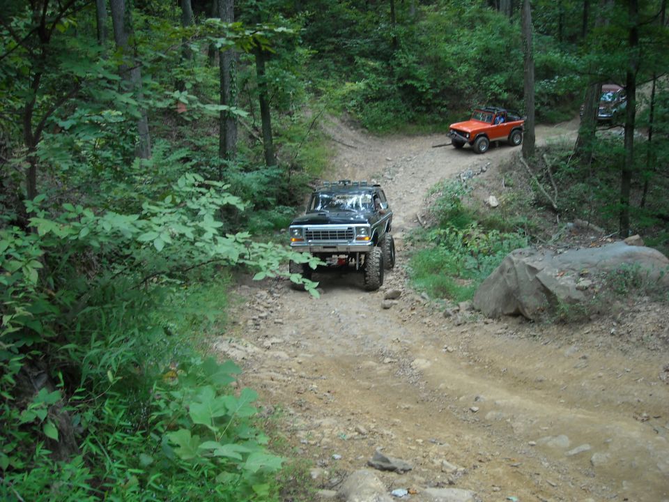 Lifted truck and orange Bronco on a wooded dirt trail with a boulder