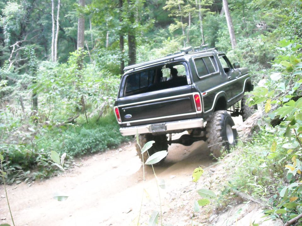 Black lifted Ford Bronco climbing a rutted forest trail from the rear