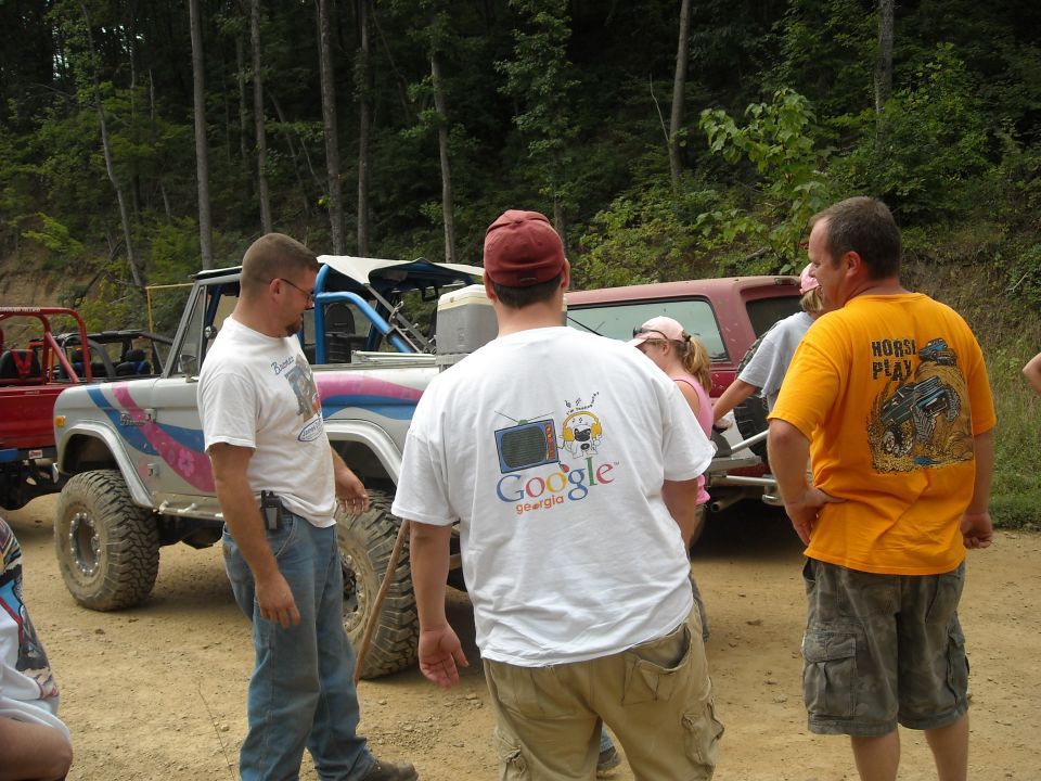 Group of men in t-shirts standing around lifted trucks at a trail clearing