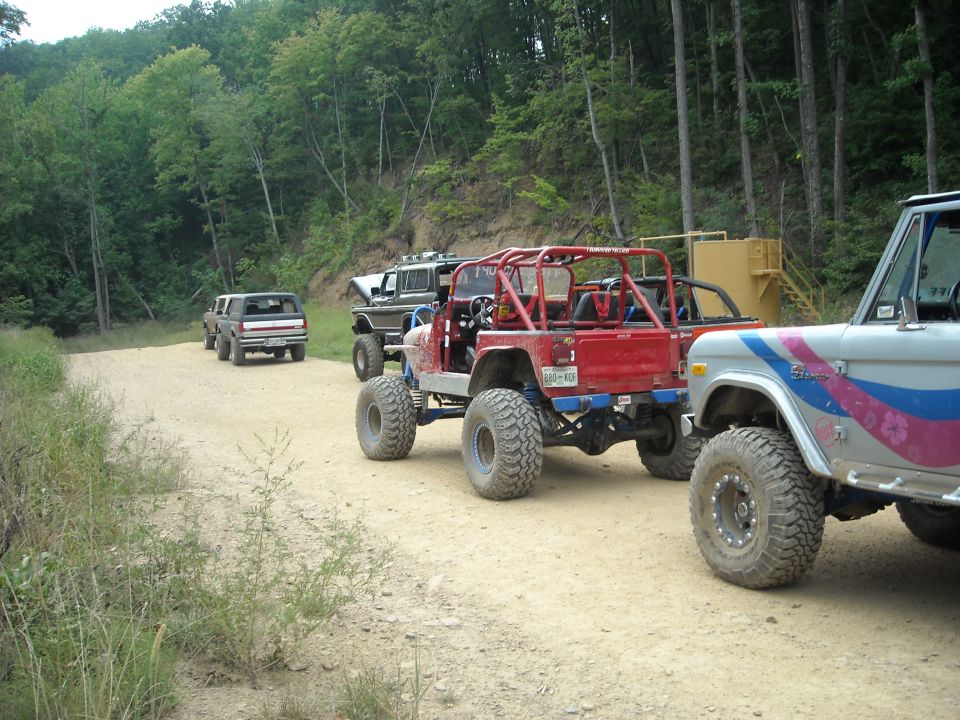 Line of lifted off-road trucks and a Jeep parked along a wooded dirt trail