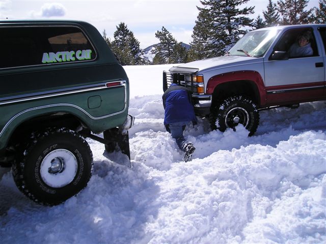Person in snow working on a stuck pickup truck next to a green SUV.