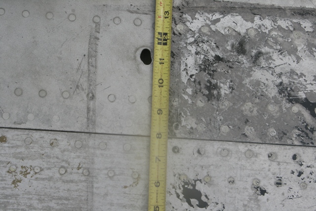 Close-up of a riveted metal surface with a hole and a tape measure.