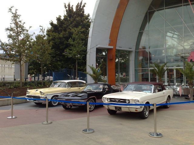 Three classic cars including a white Mustang on display outside a modern building