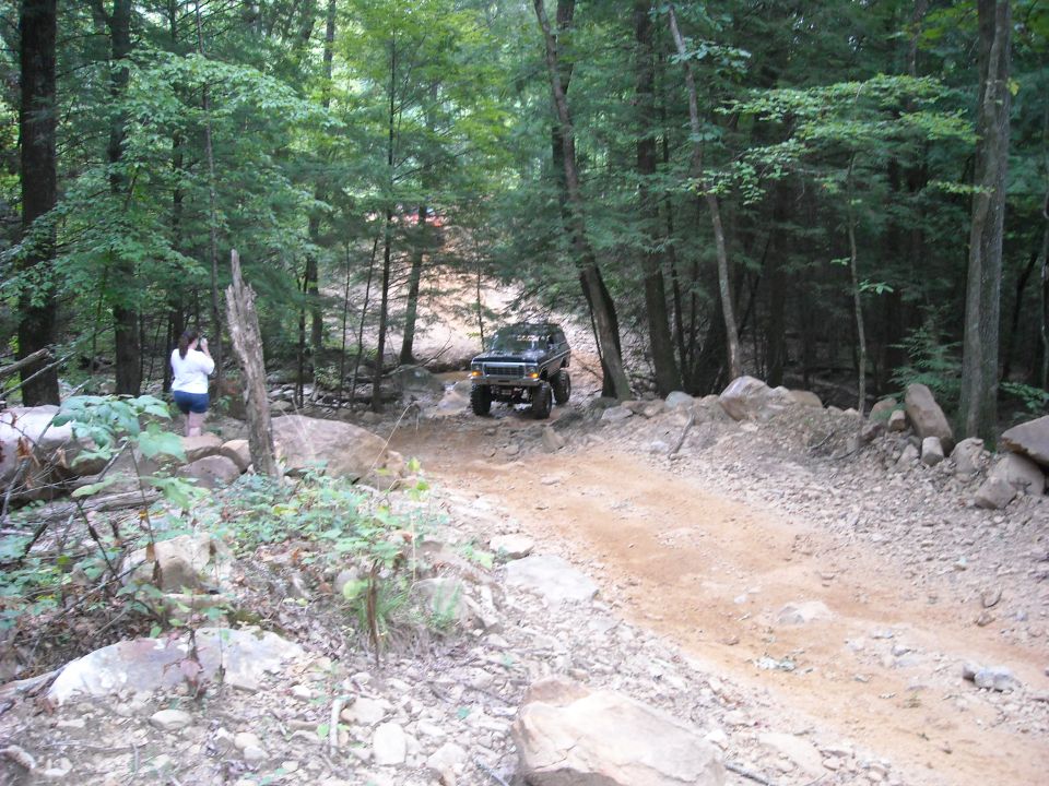 Off-road vehicle on a dirt trail in dense woods with a person watching