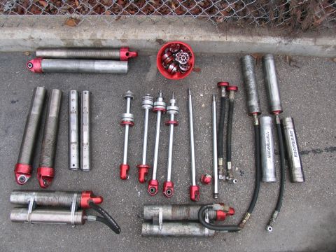 1984 Ford Bronco - A collection of various shock absorber parts, including cylinders, shafts, and hoses, are laid out on a paved surface next to a chain-link…
