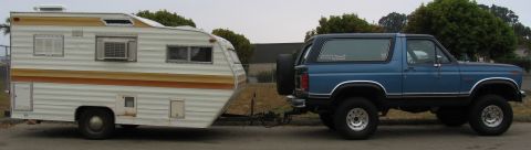 1984 Ford Bronco - A blue 1980s Ford Bronco is parked next to a white and orange striped travel trailer. The Bronco has a spare tire mounted on the back and i…