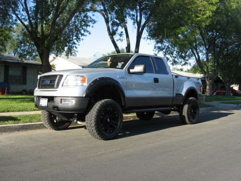 1979 Ford Bronco - A lifted, silver Ford F-150 pickup truck is parked on a residential street. The truck features large black rims and off-road tires, with a…