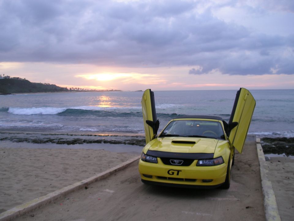 Yellow Ford Mustang with butterfly doors open on a beach at sunset.