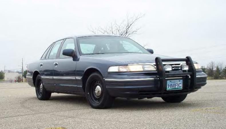 Dark blue Crown Victoria sedan with black push bar and steel wheels, front plate HMFIC, parked on empty lot