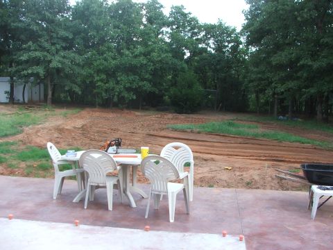 2005 Ford Mustang - A patio area with white plastic chairs and a table is set up in front of a wooded area with disturbed earth and tire tracks.