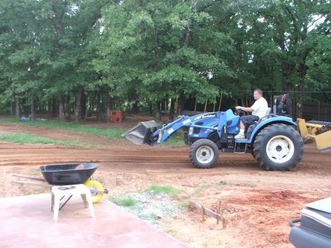 2005 Ford Mustang - A man drives a blue New Holland tractor with a front loader and a grader attachment in a dirt lot with trees in the background.