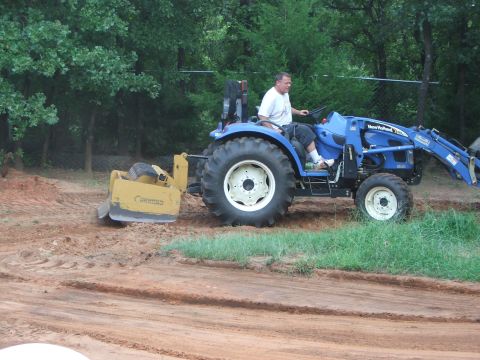 2005 Ford Mustang - A man operates a blue New Holland tractor with a yellow land plane attachment on a dirt lot. The tractor is moving dirt, and the land plane…