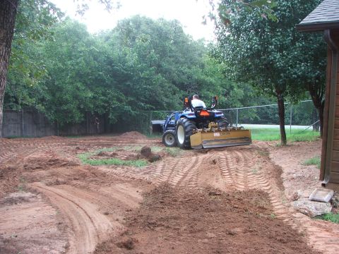 2005 Ford Mustang - A blue tractor with a yellow grader attachment is leveling a dirt area. The scene is outdoors with trees and a fence in the background.