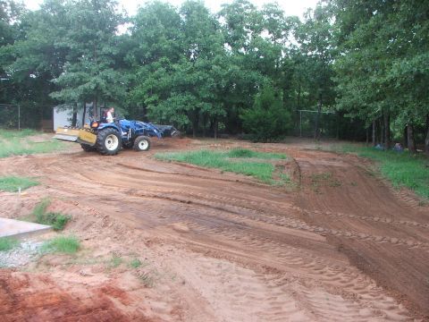 2005 Ford Mustang - A blue tractor with a person operating it is working on a dirt track. The tractor has a front loader and a rear attachment, possibly a till…