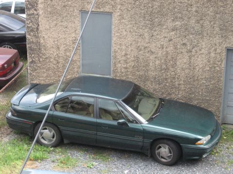 1992 Ford Crown Victoria - A dark green four-door sedan is parked on a gravel lot next to a textured concrete building. The car has a spoiler on the trunk and is view…